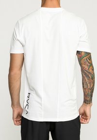 Bílé tričko s krátkým rukávem a žebrovaným kulatým výstřihem, s vertikálními švy a černým logem "RVCA" na boku. Hladká textura.