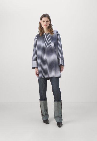 Tunique rayée bleue et blanche, présentant un design en chevrons au niveau du col, associée à un jean en denim et à des manches évasées fendues distinctives.