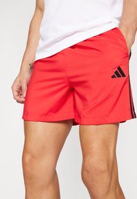 Red athletic shorts made of lightweight fabric, featuring three black stripes and an Adidas logo accent on the lower side.