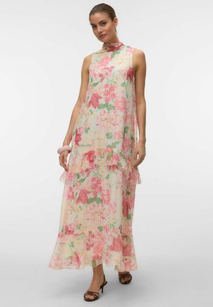 Femme portant une robe maxi sans manches à col montant superposée avec un imprimé floral rose et vert, associée à des sandales marron à talons et un bracelet.