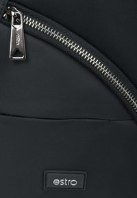 Czarno tkaninowy plecak w zbliżeniu, z metalowym zamkiem błyskawicznym w kolorze srebrnym oraz prostokątną naszywką z logo, na której widnieje nazwa "estro". Gładka tekstura.
