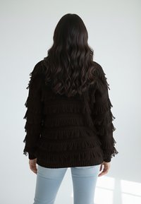 Brązowy, dziergany sweter z frędzlami, ozdobiony teksturowanymi pasami, z długimi rękawami i luźnym krojem, w połączeniu z dopasowanymi jasnoniebieskimi jeansami.