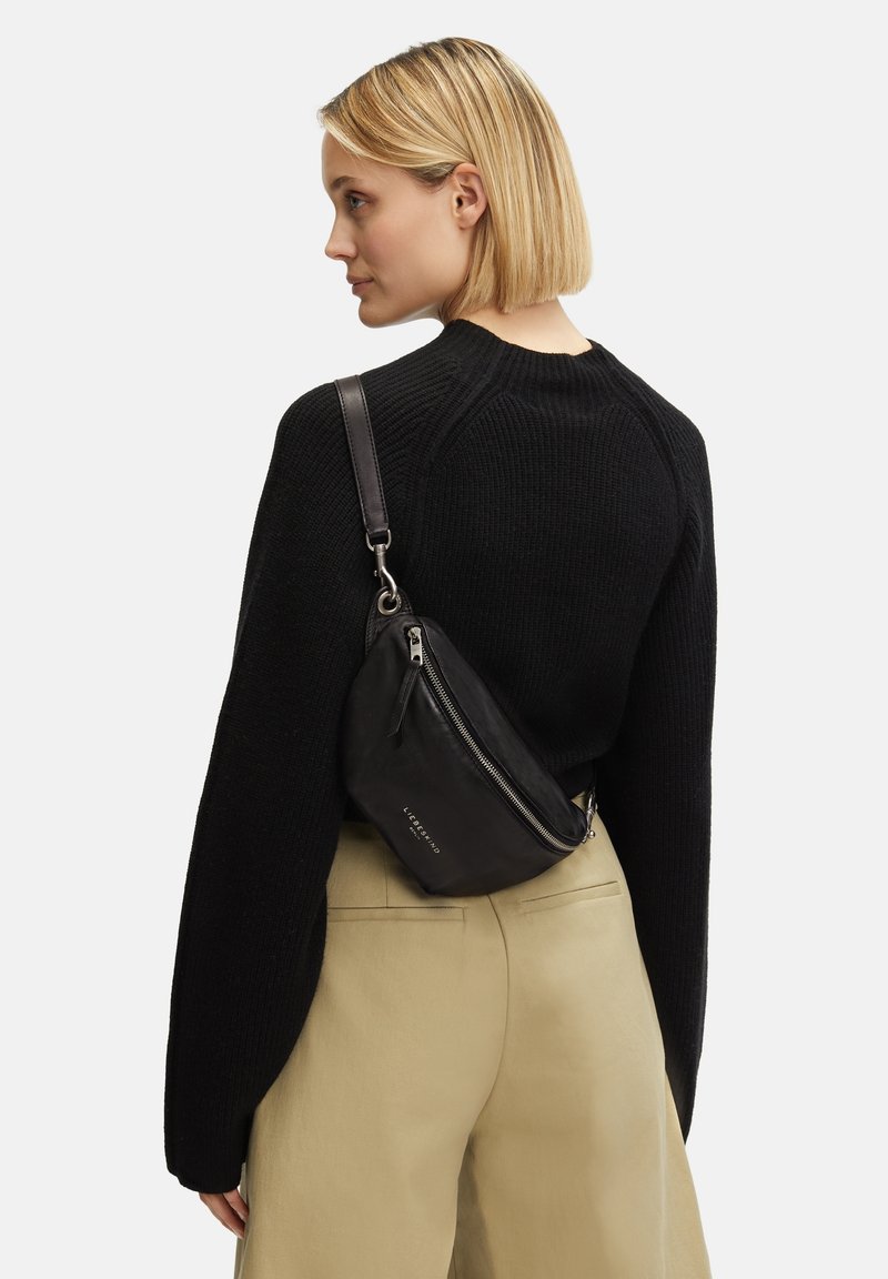 Borsa a spalla nera realizzata in morbida pelle, con una forma curva, chiusura con zip e dettagli in metallo argentato. Indossata su un pullover nero a maglia.