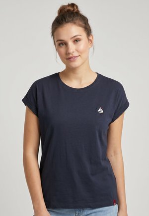 Junge Frau mit Haaren im Dutt, die ein dunkelblaues T-Shirt mit hochgekrempelten Ärmeln und einem kleinen Segelboot-Logo auf der Brust trägt, steht vor einem schlichten Hintergrund.