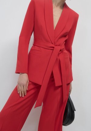 Kvinde iført en skarpt rød blazer med bælte og matchende vidde bukser, der holder en sort håndtaske mod en ensfarvet baggrund.