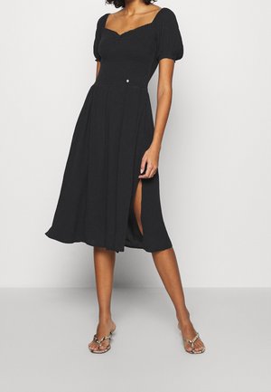 Frau in einem schwarzen Midikleid mit Puffärmeln und Seitenschlitz, trägt Sandalen mit Schlangenmuster und offenen Zehen, steht vor einem schlichten Hintergrund.