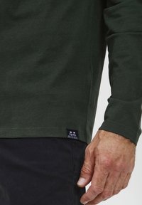 Langærmet mørkegrøn T-shirt lavet af bomuld, med en lige kant og et lille mærke nederst til venstre.