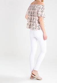 Blusa off-the-shoulder in rosa chiaro con motivo floreale, abbinata a jeans skinny bianchi e sandali slip-on in tonalità neutre.