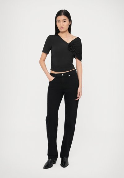 Čierny top s krátkym rukávom a asymetrickým výstrihom s kvetinovým akcentom, skombinovaný s čiernymi rovnými džínsami a čiernymi členkovými topánkami so špicatou špičkou.