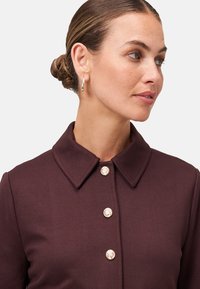 Robe bourgogne à col avec un design structuré, ornée de boutons dorés proéminents et d'une texture lisse. Le modèle porte des boucles d'oreilles créoles.
