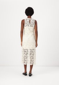 Crème-kantjurk met hoge hals en uitgesneden achterkant, versierd met bloemenpatronen en een lengte tot halverwege het kuit. Draagt met zwarte instappers.