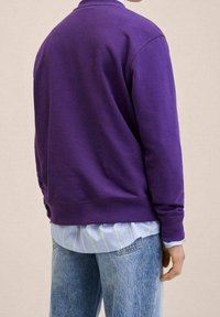 Sudadera de forro polar morada con cuello redondo y puños acanalados, encima de una camisa a rayas azul claro, combinada con jeans azul claro.