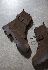 Brązowe zamszowe buty bojowe z grubymi podeszwami z wypustkami, sznurówkami z przodu, paskami na klamry i pętlami do zakupu na szarej teksturowanej powierzchni.