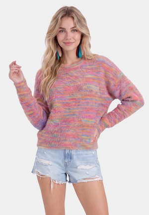 Junge Frau mit langen blonden Haaren, die einen bunten, flauschigen Pullover, große mehrfarbige Federohrringe und ausgewaschene helle Jeansshorts trägt und lächelt.