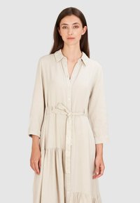 Młoda kobieta z długimi brązowymi włosami, ubrana w beżową sukienkę zapinaną na guziki z paskiem w talii i rękawami o długości 3/4, stojąca na jednolitym tle.