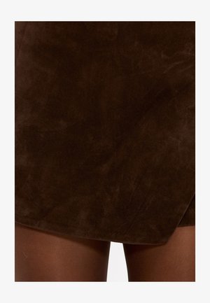 Mini-jupe en daim marron avec un léger effet portefeuille, présentant une texture lisse et des bords nets. Assortie à des collants transparents marron.