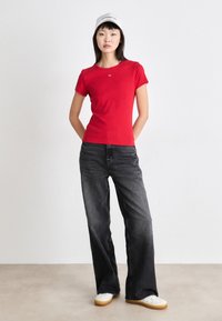 Camiseta roja de manga corta, diseño ajustado. Combinada con unos vaqueros de pierna ancha negros desgastados y zapatillas blancas. El modelo lleva una gorra blanca.
