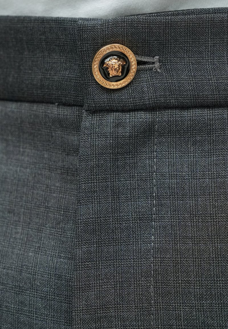 Grijs gestructureerd stof met fijne ruitpatronen, met een gouden en zwarte ronde knoop als accent, met een Medusa-logo in het midden.