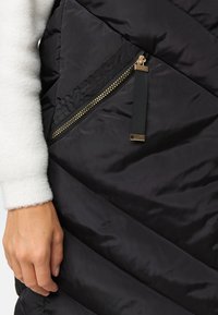 Veste matelassée noire avec coutures diagonales et poche zippée dorée avec languette en tissu ; manchette de pull blanc doux visible.