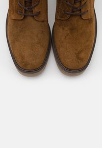 Brązowe zamszowe botki za kostkę z zaokrąglonymi noskami, ciemnobrązowymi sznurowadłami i delikatną gumową podeszwą. Gładka faktura i minimalistyczne detale wzornicze.