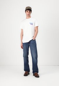 Hvid t-shirt med multicolor grafik, blå denim jeans, brune slip-on sko og en brun ternet kasket. Casual outfit med en afslappet pasform.