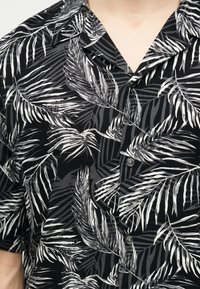Μαύρο πουκάμισο με κοντά μανίκια και κουμπιά, με λευκό και γκρι τροπικό σχέδιο φύλλων φοίνικα, που φοριέται από άτομο με ανοιχτόχρωμο δέρμα ορατό στον λαιμό.