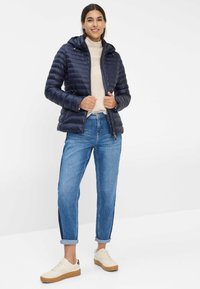 Marine blauer Fleecejacke mit Kapuze, Reißverschluss vorne und horizontalen Nähten; kombiniert mit hellblauen Jeans und beigen Sneakers mit Gummisohlen.