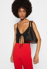 Žena s přirozeně kudrnatými vlasy, oblečená v černém krátkém topu se zavazováním vpředu, červených kalhotách s vysokým pasem, zlatých kruhových náušnicích a s černou kabelkou přes rameno.