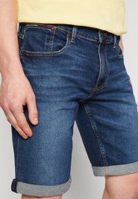 Close-up of person wearing rolled-up dark blue denim shorts and yellow shirt, with hand resting alongside the left thigh.