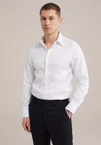 Camisa blanca de manga larga con cuello clásico y botón al frente, combinada con pantalones oscuros, destacando una textura suave y un diseño entallado.