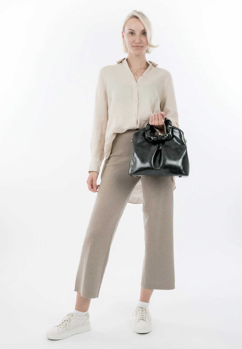Black handbag with a gathered top handle, glossy texture, and a rounded body, paired with light-colored linen shirt and beige trousers.