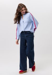 Hellblau gestreiftes Hemd mit Knöpfen und roten Seiteneinsätzen, kombiniert mit weit geschnittenen dunklen Denim-Jeans und rotem Schuhwerk. Glatte Stoffstruktur.