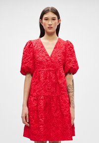 Raudona gėlėmis siuvinėta suknelė su trumpomis pūstomis rankovėmis, V formos iškirpte ir sluoksniuotais kraštais. Tekstūruotas audinys suteikia vizualinį gilumą.