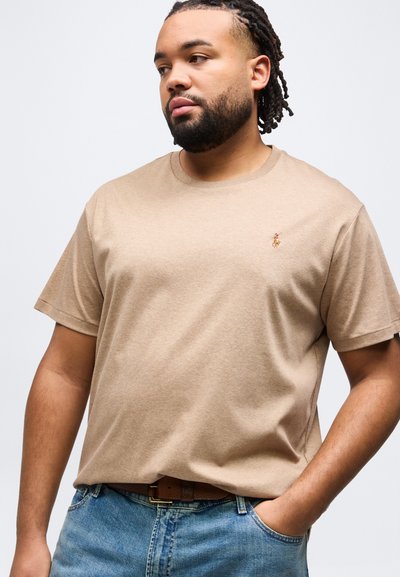 Άνδρας με γένια και πλεγμένα μαλλιά φοράει μπεζ κοντομάνικη μπλούζα με μικρό κεντημένο λογότυπο, μπλε τζιν και καφέ ζώνη, με το χέρι στην τσέπη.