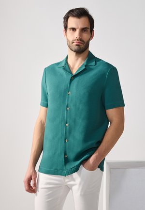 Chemise aqua à manches courtes avec un motif texturé, col structuré, fermeture à boutons et boutons dorés, associée à un pantalon blanc.