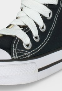 Gros plan d'une basket en toile noire avec semelle en caoutchouc blanc, lacets blancs passés dans des œillets métalliques et couture blanche distinctive.