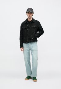 Μαύρο τζιν μπουφάν με μπροστινές τσέπες, ανοιχτό μπλε τζιν παντελόνι με ίσια γραμμή και πράσινα αθλητικά παπούτσια. Το μοντέλο φοράει γκρι καπέλο με λογότυπο.