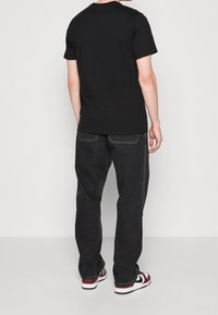 T-shirt nera a maniche corte in cotone, abbinata a jeans scuri. La vista mostra il retro dell'outfit e delle scarpe da ginnastica con dettagli rossi e bianchi.