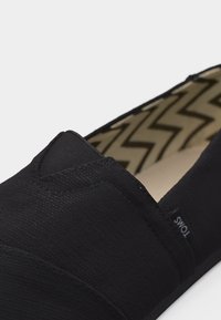 Chaussure noire en toile à enfiler avec semelle intérieure à motif zigzag et petite étiquette de marque en tissu sur le côté.