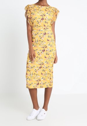 Kobieta w żółtej sukience midi w kwiaty z falbanowymi krótkimi rękawkami, nosząca białe trampki, stojąca na jednolitym białym tle.