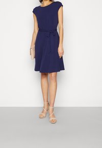 Marinblå klänning med kort ärm och midjeband, A-linje-siluett, mjukt tyg och veck. Bärs med beige sandaletter med remmar och en kuvertväska.