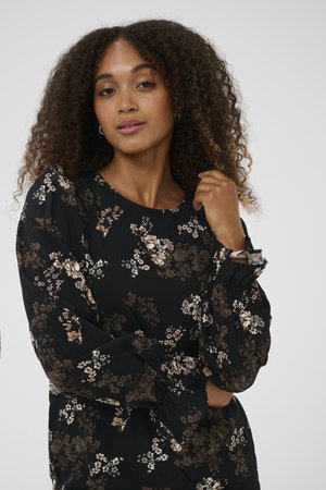 Žena s kudrnatými vlasy, oblečená v černé květinové blůze s dlouhým rukávem, pózující s jednou rukou blízko obličeje a druhou přes tělo.