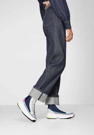 Personne portant un jean en denim foncé avec revers et des baskets montantes multicolores marchant sur un fond gris.