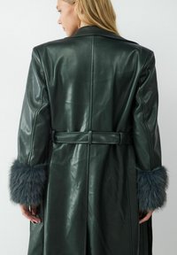 Ciemnozielony skórzany płaszcz z paskiem, z szarym futrzanym wykończeniem na rękawach i gładką fakturą. Klasyczny kołnierz i dopasowany fason.