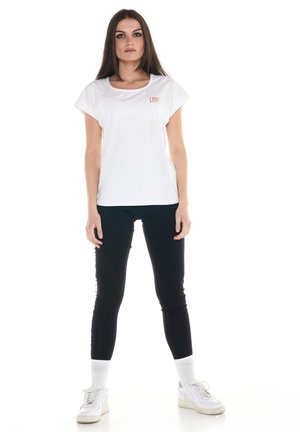Une femme porte un T-shirt blanc, un legging noir, des chaussettes blanches et des baskets blanches sur un fond blanc uni.