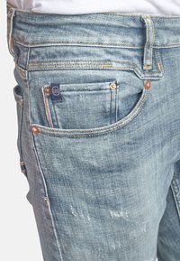 Jean en denim bleu clair avec une texture délavée, doté de plusieurs poches, de ferrures en cuivre et d'un patch logo sur la poche arrière.