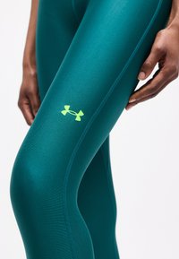 Κοντινό πλάνο σε αθλητικό κολάν πετρόλ με φθορίζον λογότυπο στον μηρό και ένα χέρι που ακουμπά κοντά στο ισχίο, σε λευκό φόντο.