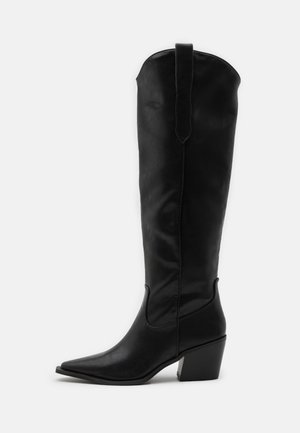 Botte de cowboy haute en cuir noir avec bout pointu et talon incliné de hauteur moyenne sur fond blanc.