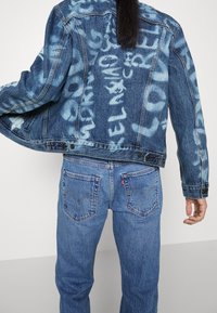 Blaue Jeansjacke mit hellblauen Graffiti-Mustern, im klassischen Schnitt, mit Knopfverschluss und zwei Fronttaschen. Kombiniert mit passenden Jeans.