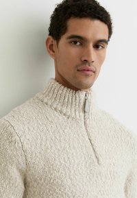 Kremno obarvan pulover z grobo pletenino, visokim ovratnikom in polovico zadrgo. Teksturirana tkanina z mehkim, udobnim videzom.